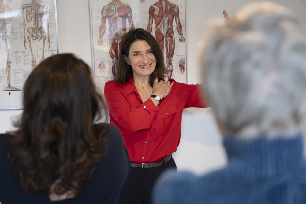 Tanya Shoop, Alexander Technique teacher, demonstrating postural awareness to students. Tanya has one arm raised with the other hand on her shoulder.